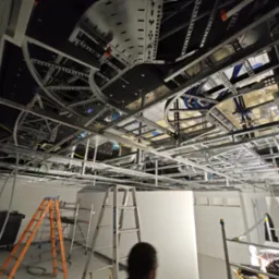 Montaż instalacji pod sufitem podwieszanym: widoczne metalowe profile, kable, rury wentylacyjne, drabiny i pracownik w tle.