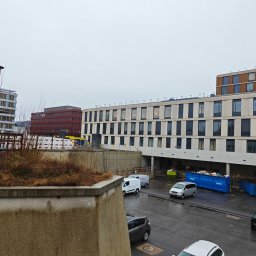 Tomasz Flis - Widok z góry na plac budowy z kontenerami, zaparkowanymi samochodami i częściowo ukończonym budynkiem z oknami w pochmurny dzień.