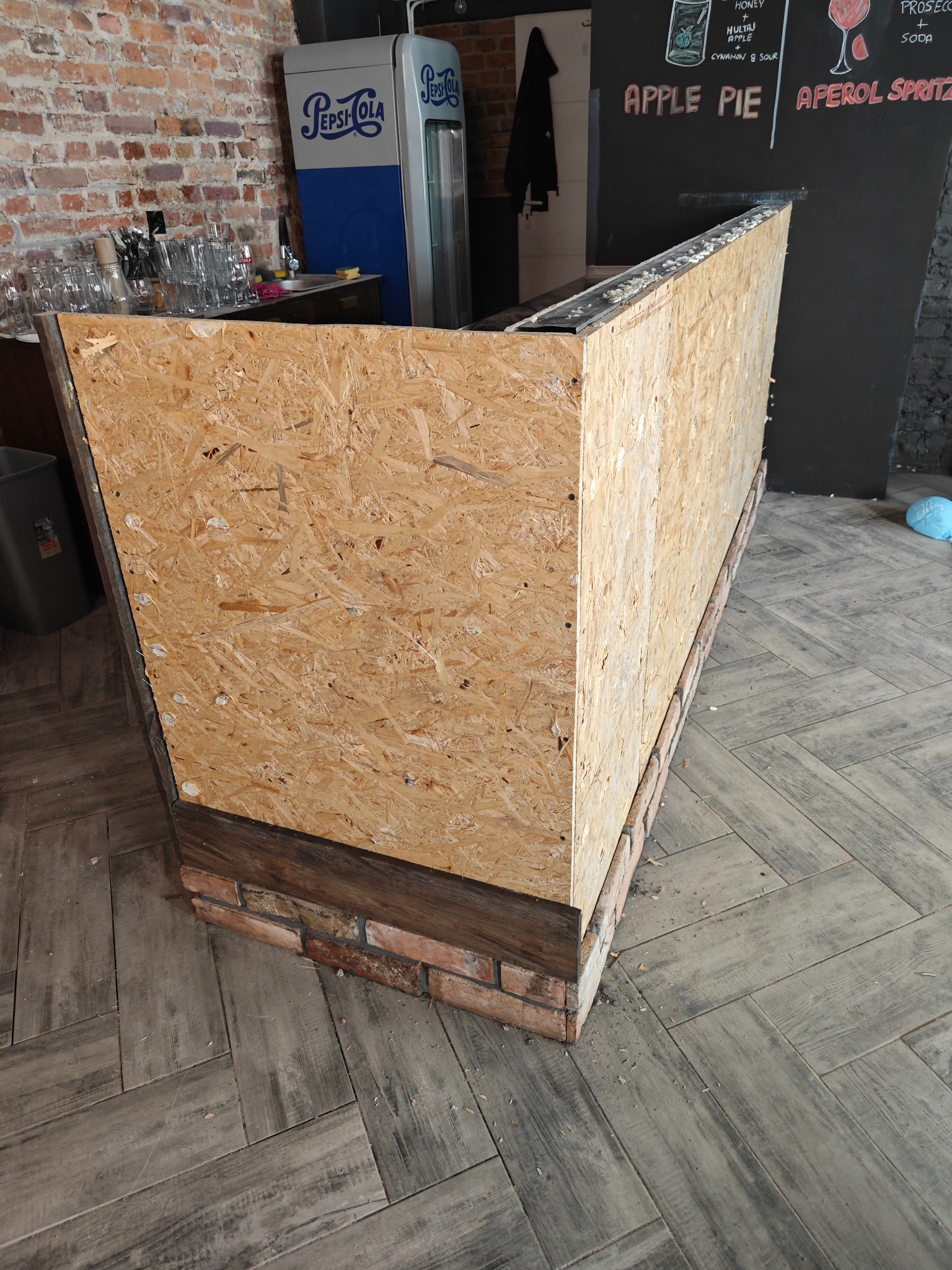 Wnętrze lokalu w trakcie remontu: surowa konstrukcja baru z płyty OSB na ceglanej podstawie, widoczne ślady po obróbce, w tle lodówka Pepsi i menu drinków na czarnej ścianie.