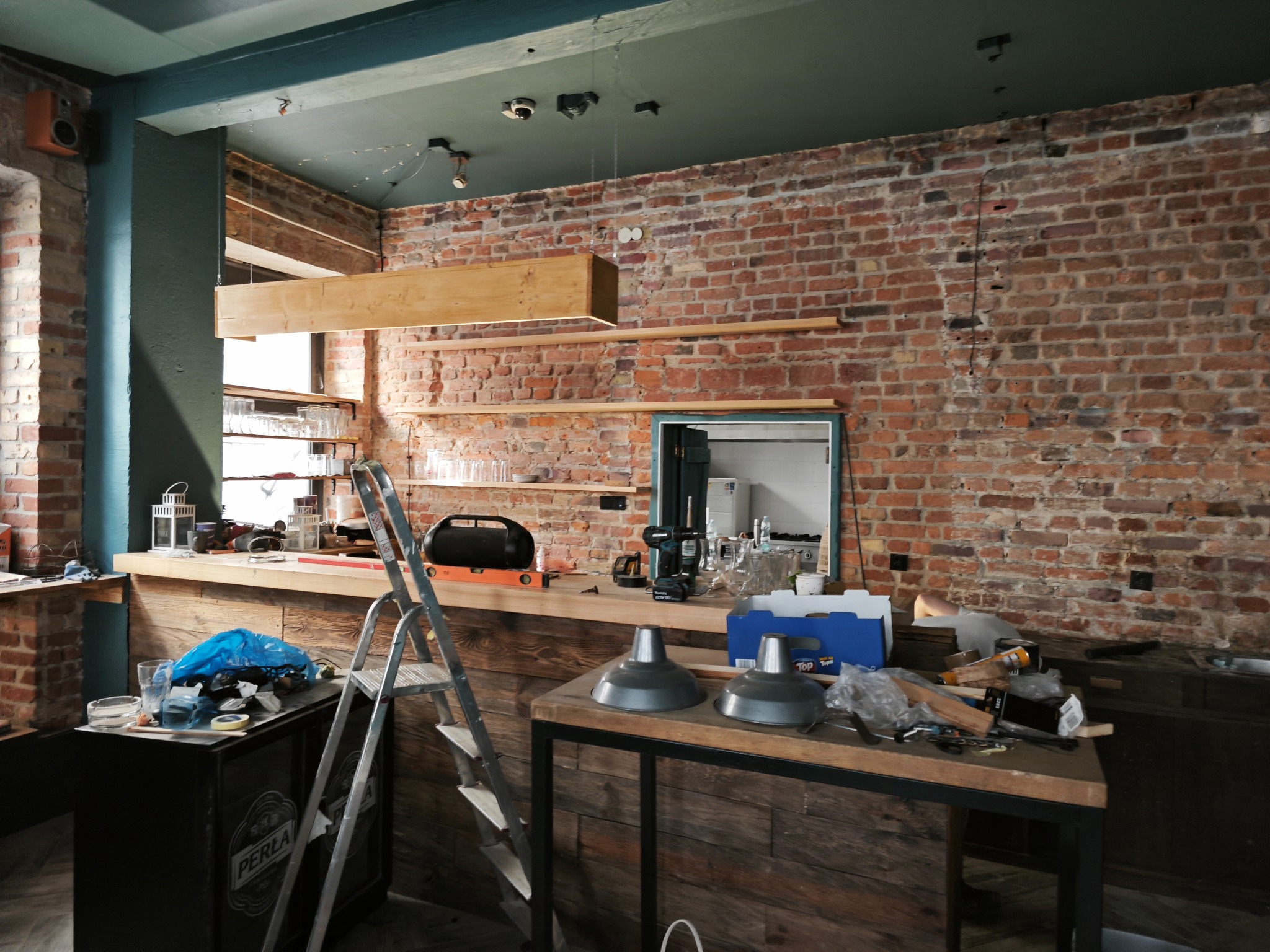 Wnętrze remontowanej restauracji z surową ceglaną ścianą, drewnianym barem w trakcie budowy, widoczne narzędzia, drabina, lampy w stylu industrialnym i elementy instalacji elektrycznej na suficie.
