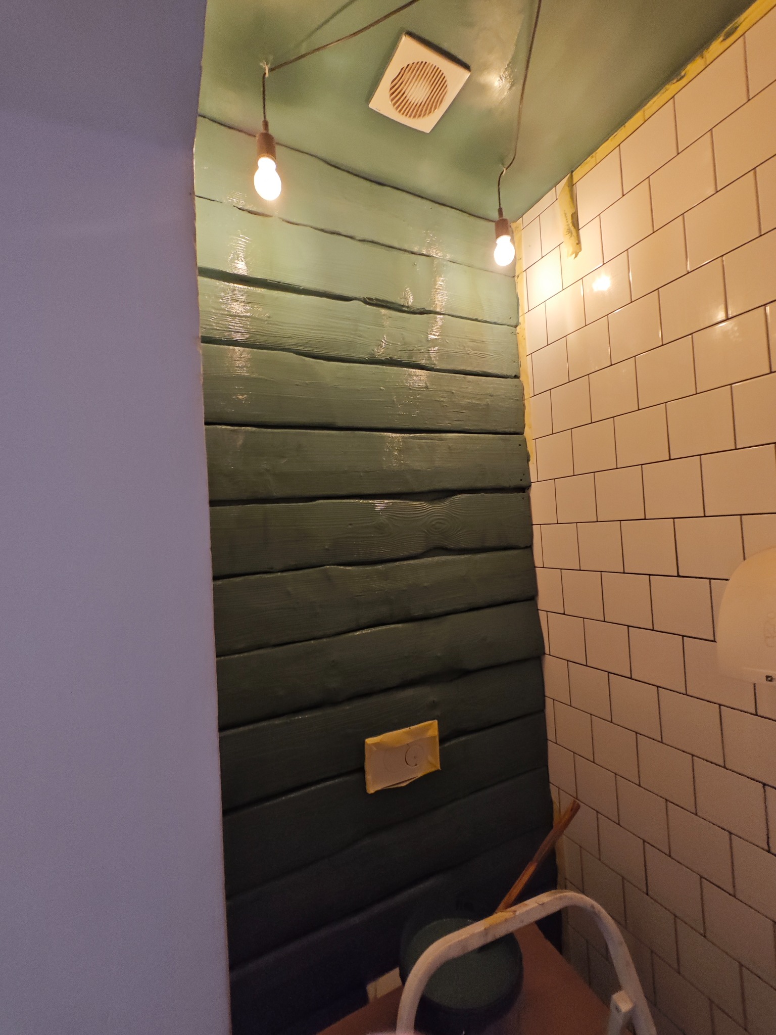 Kątowe ujęcie rogu pomieszczenia z zieloną, drewnianą ścianą obok białych płytek, widoczny wentylator sufitowy, żarówki i narzędzia malarskie