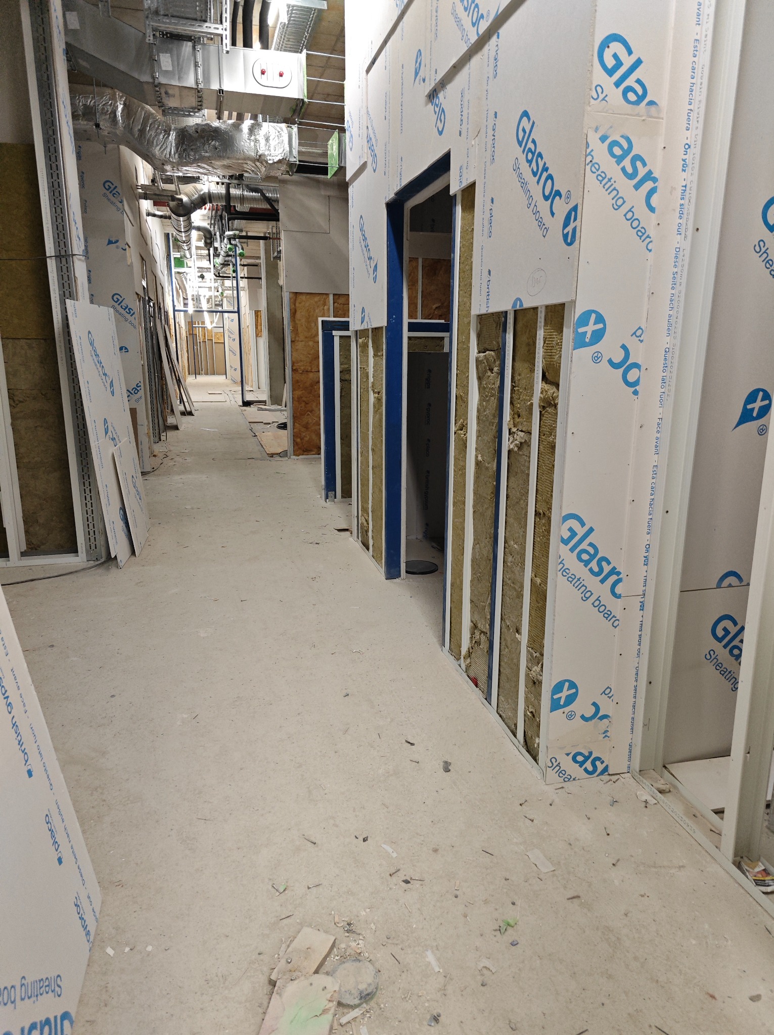 Korytarz w trakcie remontu, widoczne profile ścian działowych z wełną mineralną i częściowo zamontowane płyty Glasroc, instalacje podwieszane pod sufitem.