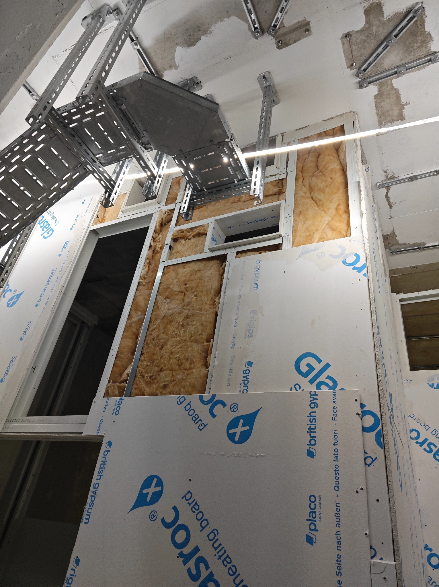 Montaż ścian działowych z płyty gipsowo-kartonowej z widoczną izolacją z wełny mineralnej, metalowe profile konstrukcyjne i koryta kablowe pod sufitem.