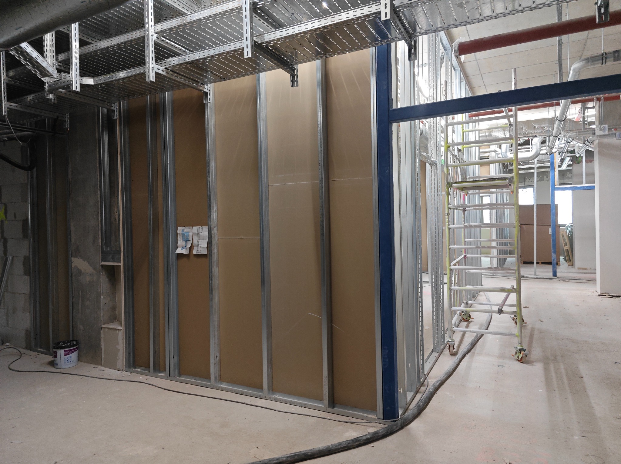 Montaż ścian działowych z płyt gipsowo-kartonowych na metalowym stelażu, widoczne instalacje pod sufitem i rusztowanie w surowym wnętrzu budynku.