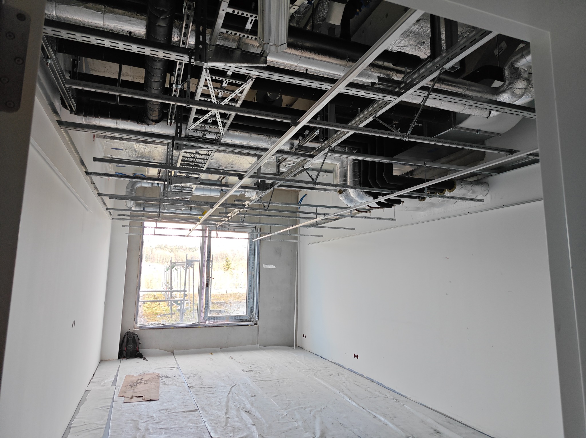 Wnętrze w trakcie remontu: widoczne metalowe profile sufitowe, instalacje wentylacyjne pod stropem, okno z widokiem na zewnątrz, podłoga zabezpieczona folią malarską, plecak w rogu pomieszczenia.