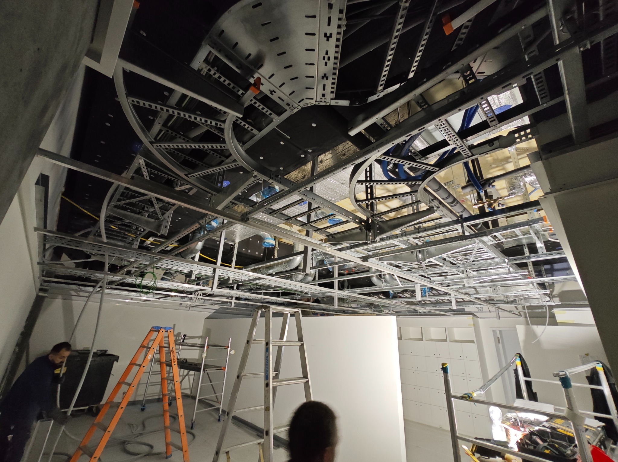 Montaż instalacji pod sufitem podwieszanym: widoczne metalowe profile, kable, rury wentylacyjne, drabiny i pracownik w tle.