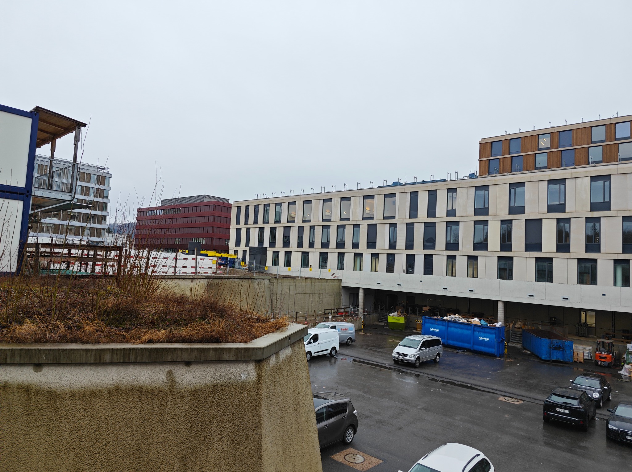 Widok z góry na plac budowy z kontenerami, zaparkowanymi samochodami i częściowo ukończonym budynkiem z oknami w pochmurny dzień.