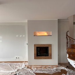 Piotr Łysik - Wnętrze w trakcie malowania, ściany w szarym odcieniu, podłoga zabezpieczona folią, widoczny kominek z kamienną obudową i dekoracyjne schody z kutą balustradą.