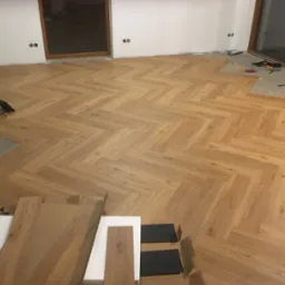 Układanie paneli podłogowych w jodełkę francuską, widoczne panele w opakowaniach i narzędzia, częściowo ułożona podłoga w jasnym odcieniu drewna.