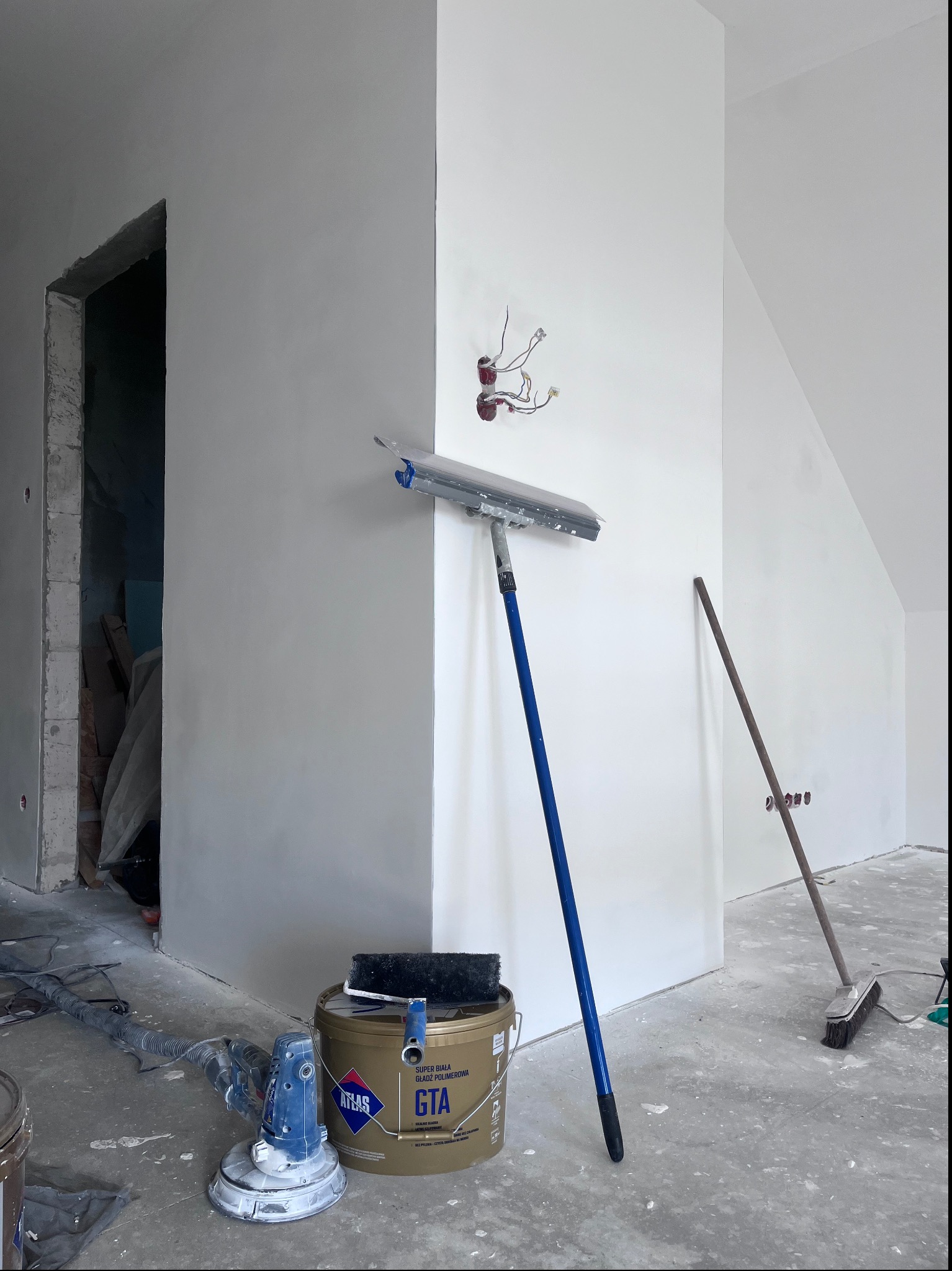 Świeżo wyszpachlowana ściana w trakcie prac wykończeniowych, widoczne narzędzia: szpachla na długim kiju oparta o ścianę, szlifierka do gładzi, wiadro z gładzią polimerową Atlas GTA i pędzel.