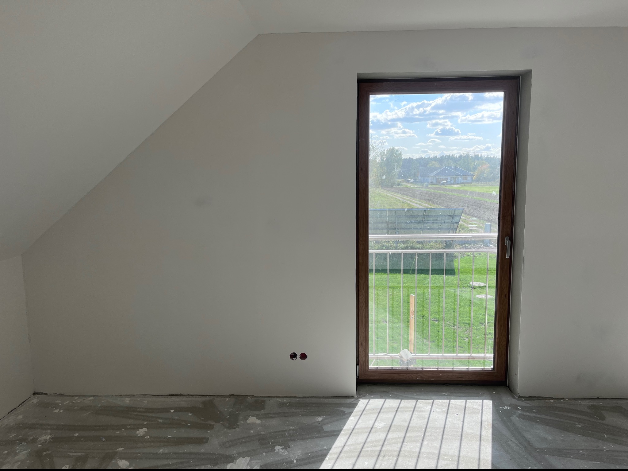 Wnętrze poddasza z oknem balkonowym, ściany po szpachlowaniu, widoczne ślady po pracach na podłodze, wystające kable elektryczne.