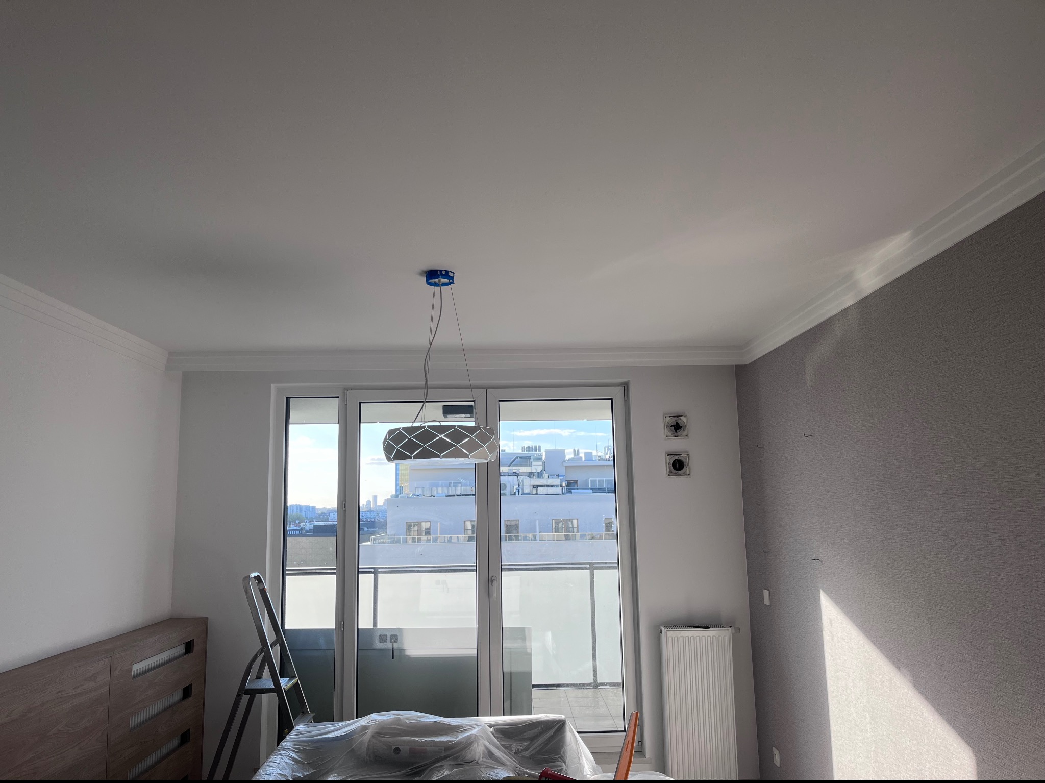 Pokój w trakcie remontu z widocznymi elementami wykończeniowymi: listwy przysufitowe, szara tapeta na jednej ścianie, sufit z przygotowanym miejscem na lampę, okno balkonowe z widokiem na miasto...