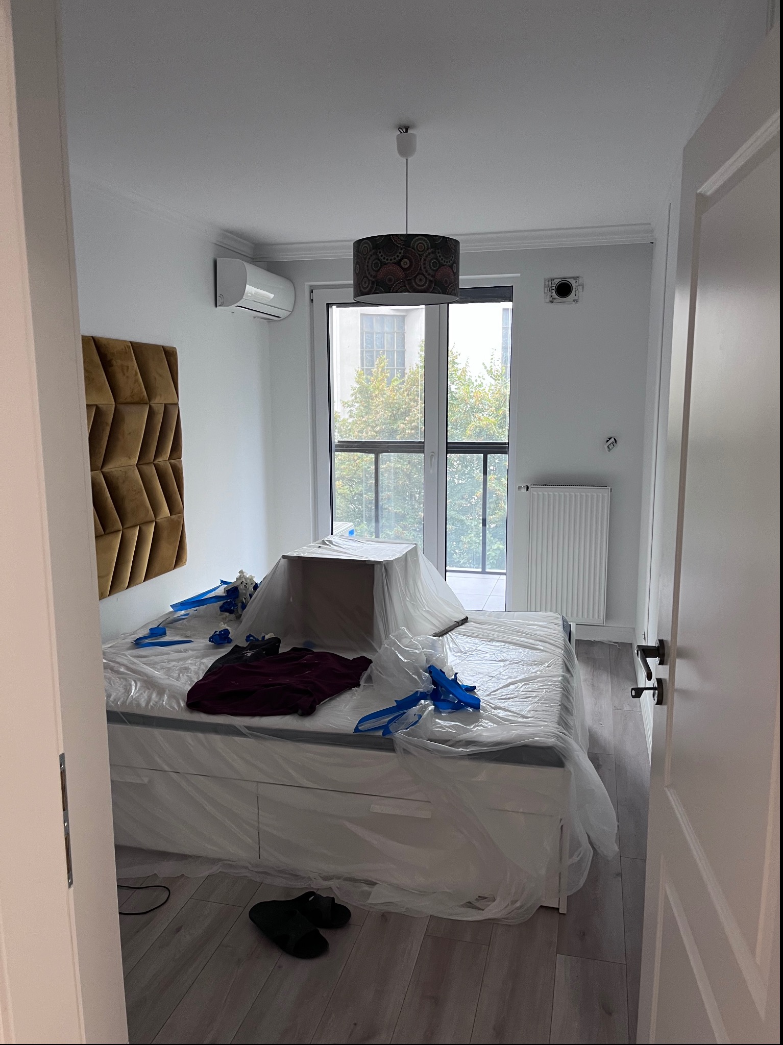 Pokój w trakcie zabezpieczania przed pracami wykończeniowymi; łóżko, meble i podłoga przykryte folią ochronną, widoczne taśmy malarskie, w tle okno balkonowe.
