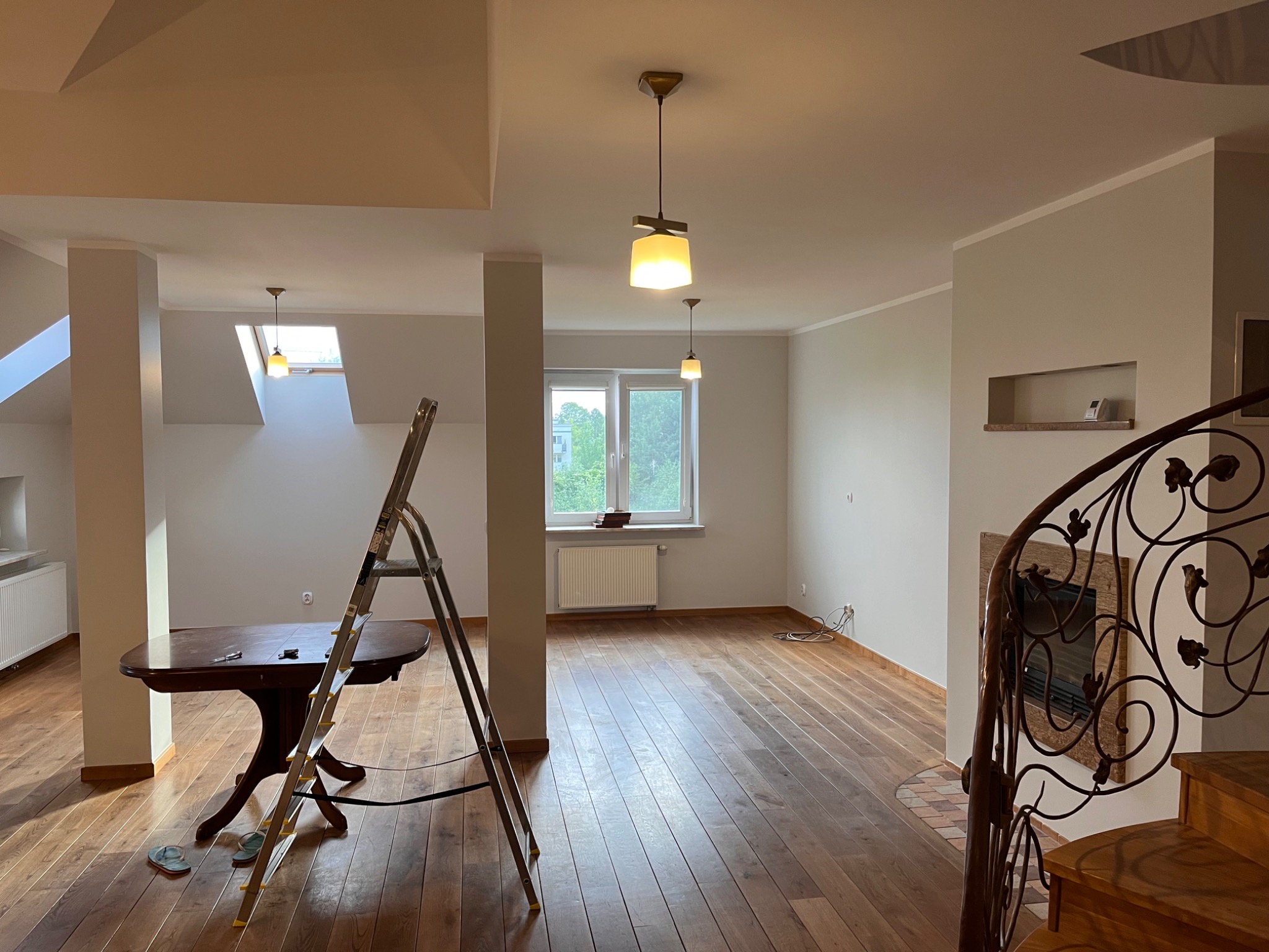 Jasne wnętrze w trakcie prac wykończeniowych, z drewnianą podłogą, schodami z kutą balustradą, aluminiową drabiną, stołem i widocznymi instalacjami elektrycznymi.