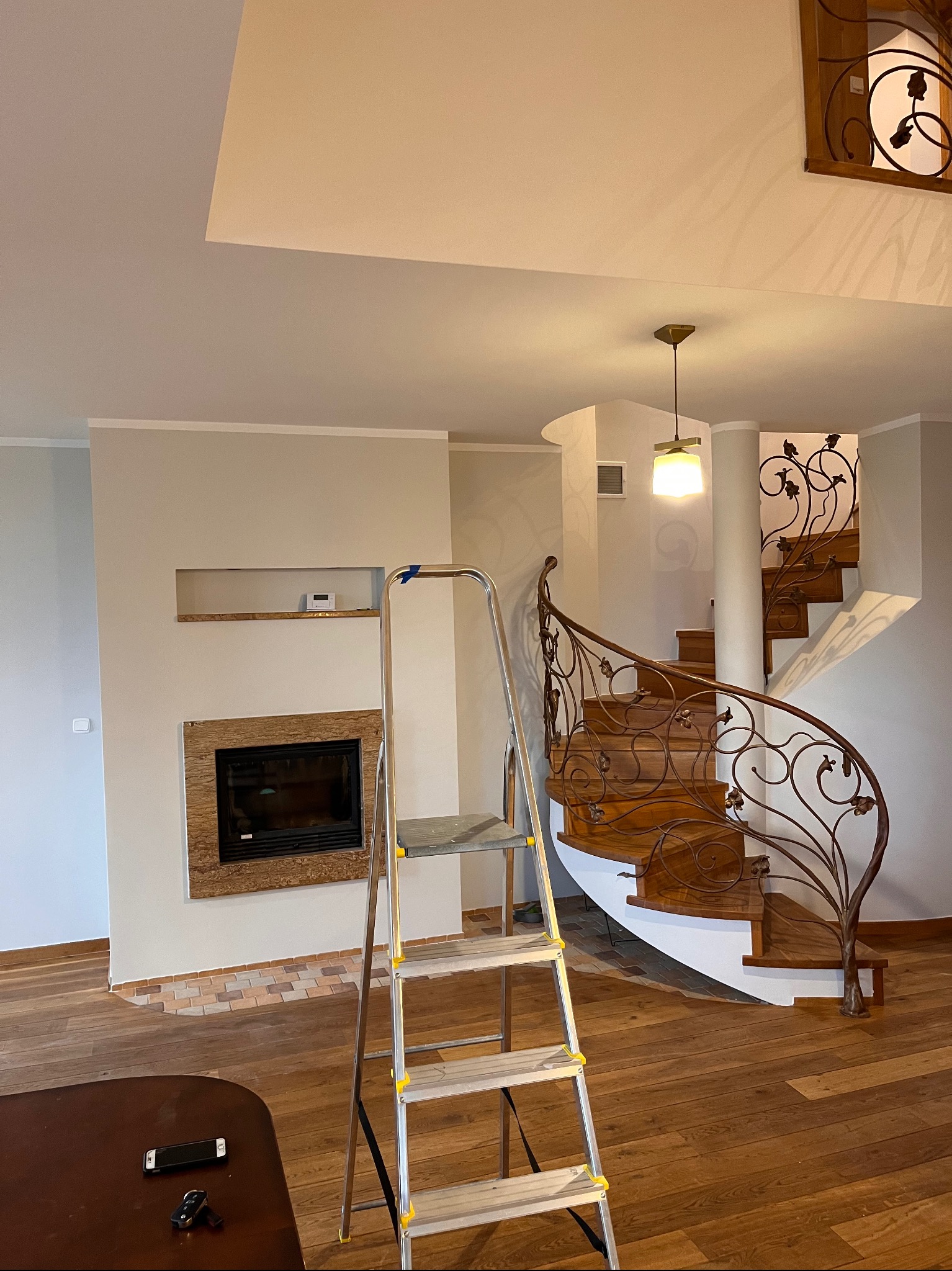 Wnętrze z drewnianymi schodami spiralnymi z kutą balustradą, kominkiem z obudową z płytek i aluminiową drabiną opartą o ścianę, prawdopodobnie w trakcie prac wykończeniowych.