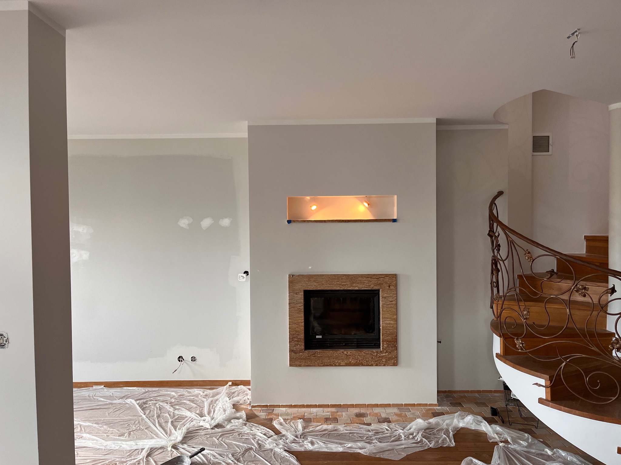 Wnętrze w trakcie malowania, ściany w szarym odcieniu, podłoga zabezpieczona folią, widoczny kominek z kamienną obudową i dekoracyjne schody z kutą balustradą.