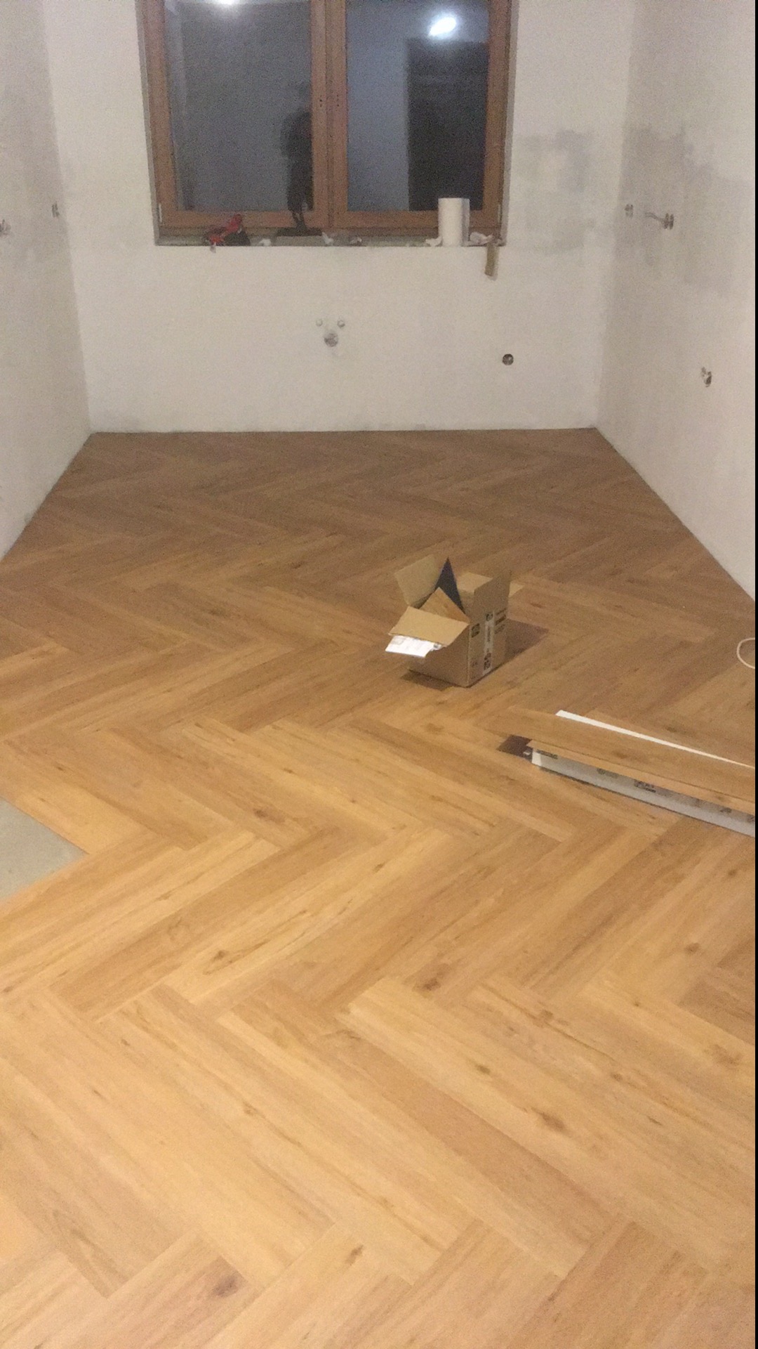 Ułożona podłoga z paneli w jodełkę w pokoju z surowymi ścianami, karton po panelach na środku, listwy przypodłogowe obok.