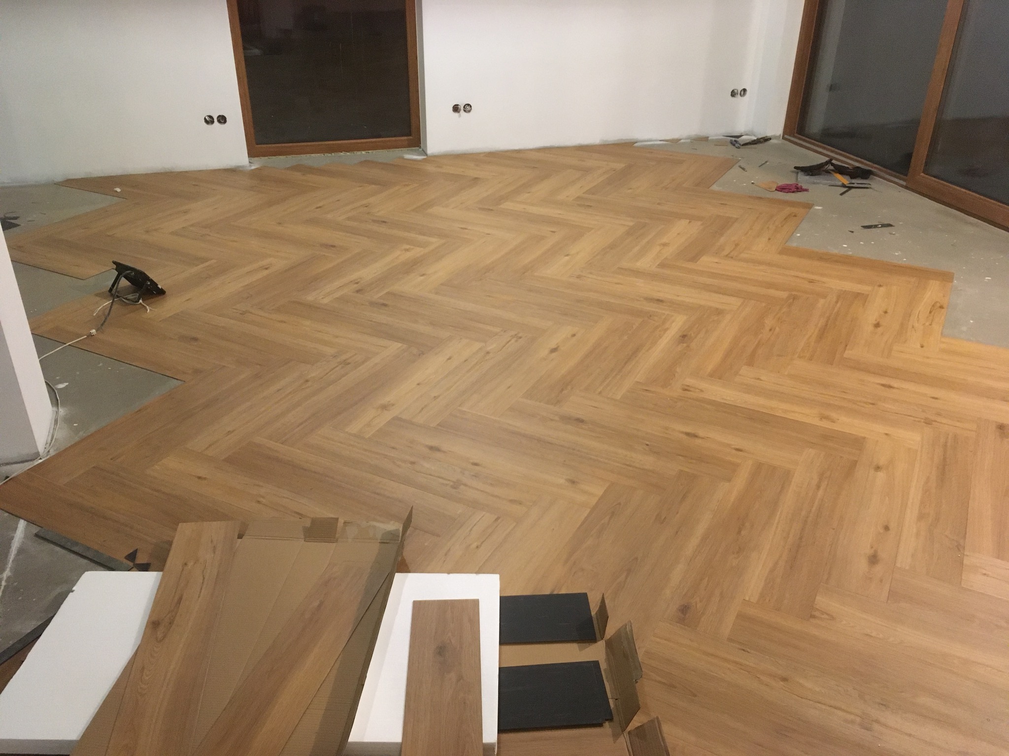 Układanie paneli podłogowych w jodełkę francuską, widoczne panele w opakowaniach i narzędzia, częściowo ułożona podłoga w jasnym odcieniu drewna.