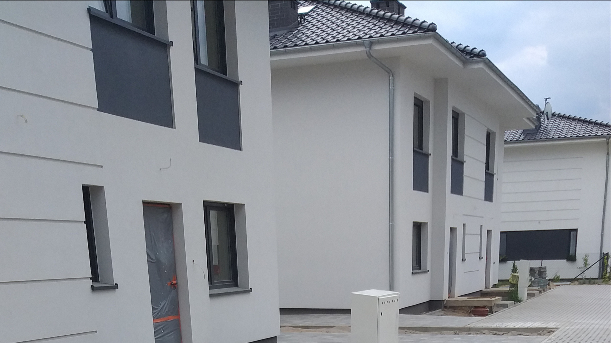 Nowoczesne domy jednorodzinne w zabudowie szeregowej, z jasną elewacją i szarymi elementami, chodnik z kostki brukowej w trakcie układania, zabezpieczone folią drzwi wejściowe.