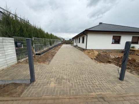 Ukończony podjazd z kostki brukowej w jodełkę prowadzący do białego domu z brązowymi okiennicami. Widoczny fragment ogrodzenia z siatki i zielone tuje.