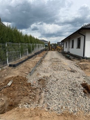Przygotowanie podłoża pod nawierzchnię: wykop, podsypka żwirowa, obrzeża. W tle koparka i dom jednorodzinny. Widok na posesję w trakcie prac ziemnych.