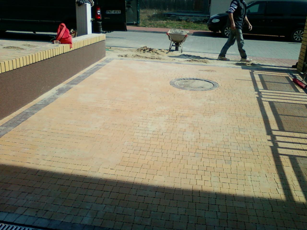 Jasna, nowo ułożona nawierzchnia z kostki brukowej z włazem kanalizacyjnym pośrodku, widoczny pracownik z taczką w tle.