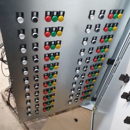 Tompor - Metalowa szafa sterownicza z rzędami białych, czarnych, czerwonych, zielonych i żółtych przycisków oraz przełącznikiem na górze, widoczne przewody po lewej stronie.