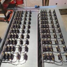 Tompor - Rozbudowana szafa sterownicza z licznymi przekaźnikami i okablowaniem, widoczne oznaczenia na przewodach, w tle elementy wyposażenia warsztatu.