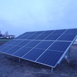 Tompor - Instalacja paneli fotowoltaicznych na gruncie, widok na rzędy ciemnoniebieskich paneli, tło: domy jednorodzinne i pochmurne niebo.