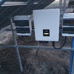 Tompor - Instalacja paneli słonecznych: widok na inwerter FOX ESS z żółtym wskaźnikiem, skrzynki przyłączeniowe i metalową konstrukcję mocującą na tle ziemi.