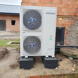 Tompor - Zewnętrzna jednostka pompy ciepła Kaisai, zamontowana na czarnych blokach fundamentowych przy ceglanej ścianie budynku, widoczny certyfikat R32 i instalacja elektryczna.