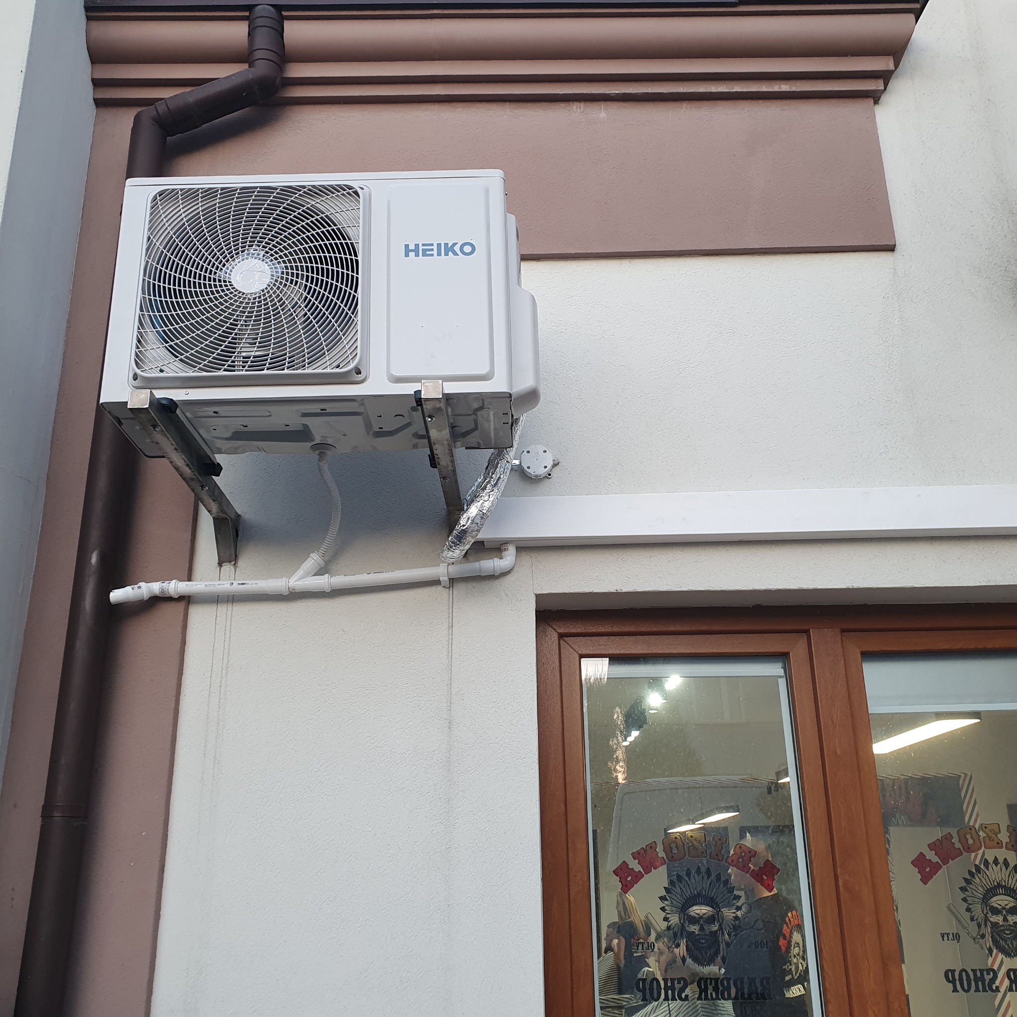 Zewnętrzna jednostka klimatyzacji marki Heiko zamontowana na ścianie budynku z brązowym orynnowaniem i oknem z odbiciem wnętrza salonu fryzjerskiego.