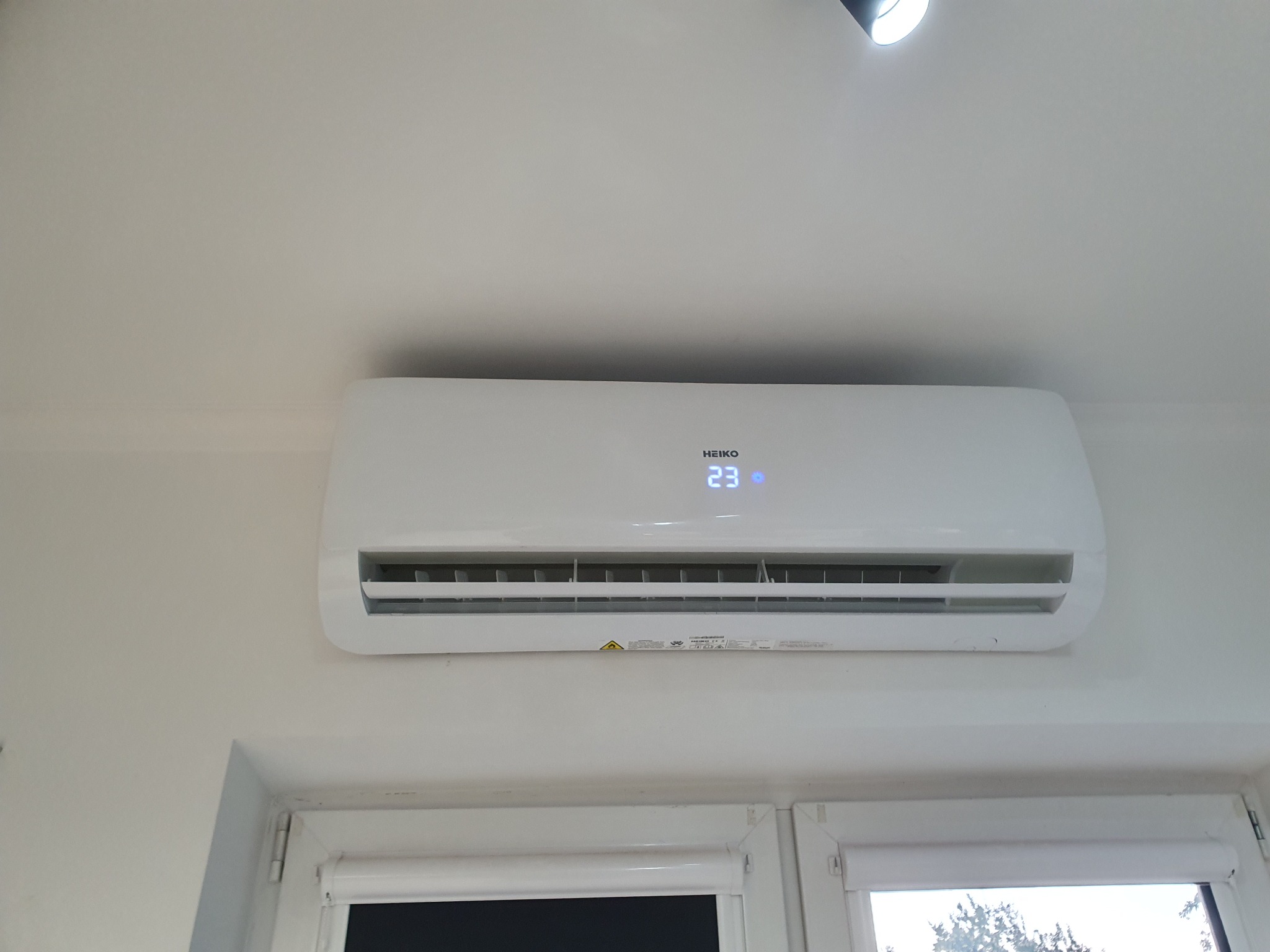 Biały klimatyzator HEIKO zamontowany na ścianie nad oknem, wyświetla temperaturę 23 stopnie Celsjusza.