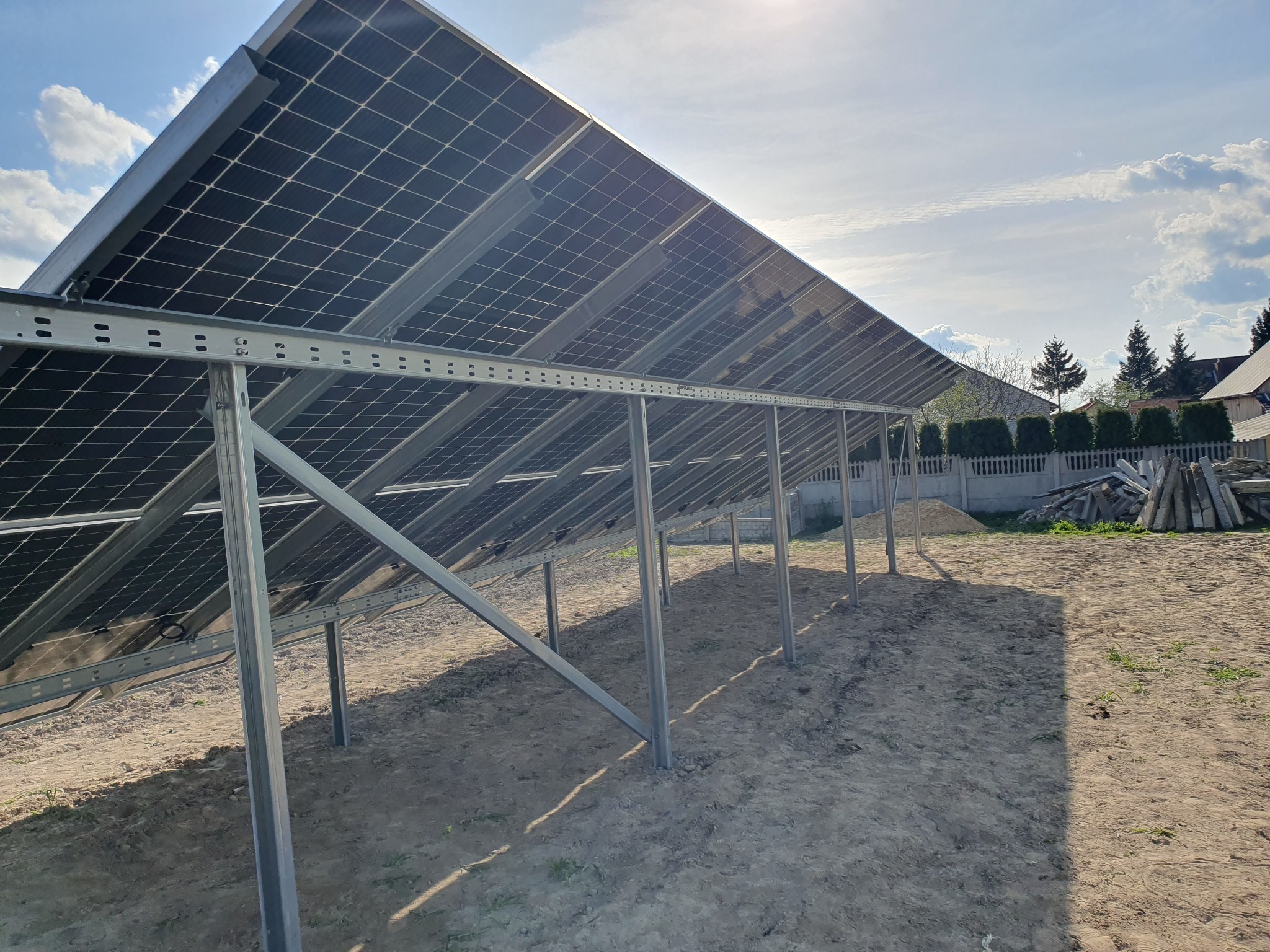 Nisko kątowe ujęcie od spodu na zamontowane panele słoneczne na metalowej konstrukcji, widoczne ślady po wierceniu w ziemi, w tle dom jednorodzinny i błękitne niebo z chmurami.