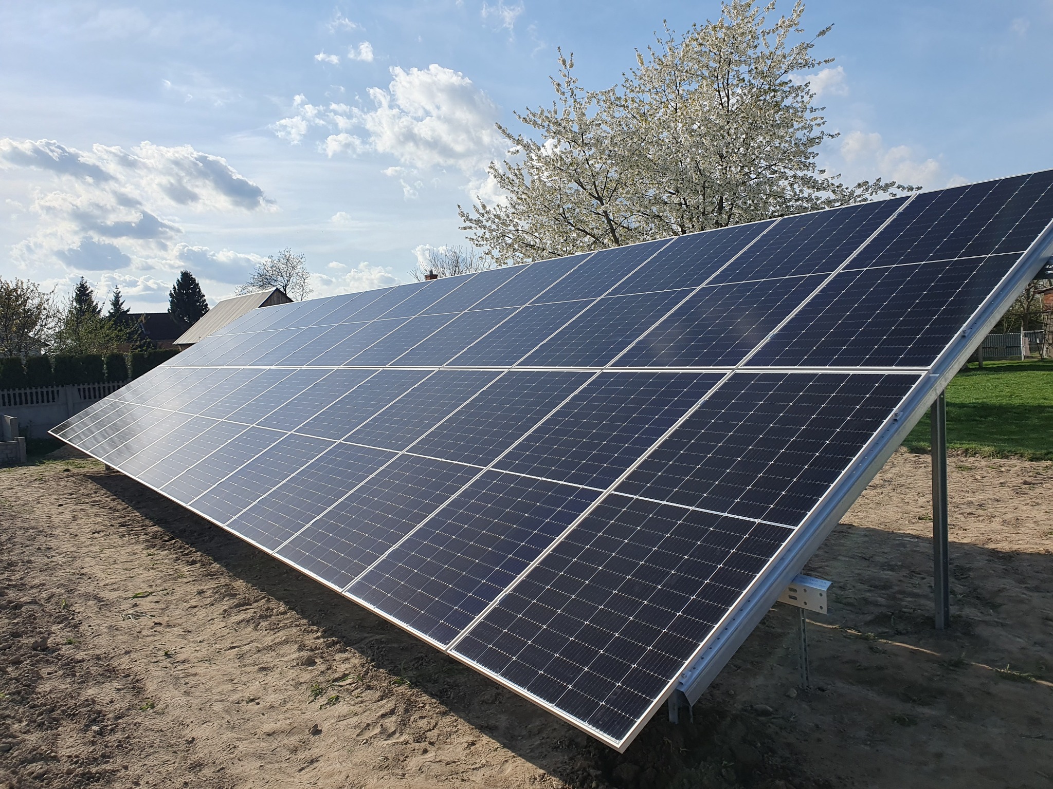 Panel słoneczny na metalowej konstrukcji, z kwitnącym drzewem i błękitnym niebem w tle, na przygotowanym podłożu.