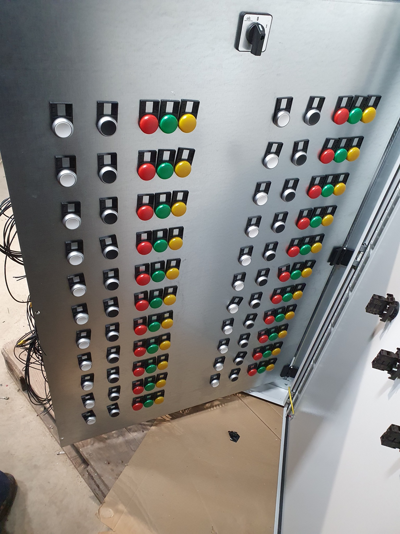 Metalowa szafa sterownicza z rzędami białych, czarnych, czerwonych, zielonych i żółtych przycisków oraz przełącznikiem na górze, widoczne przewody po lewej stronie.