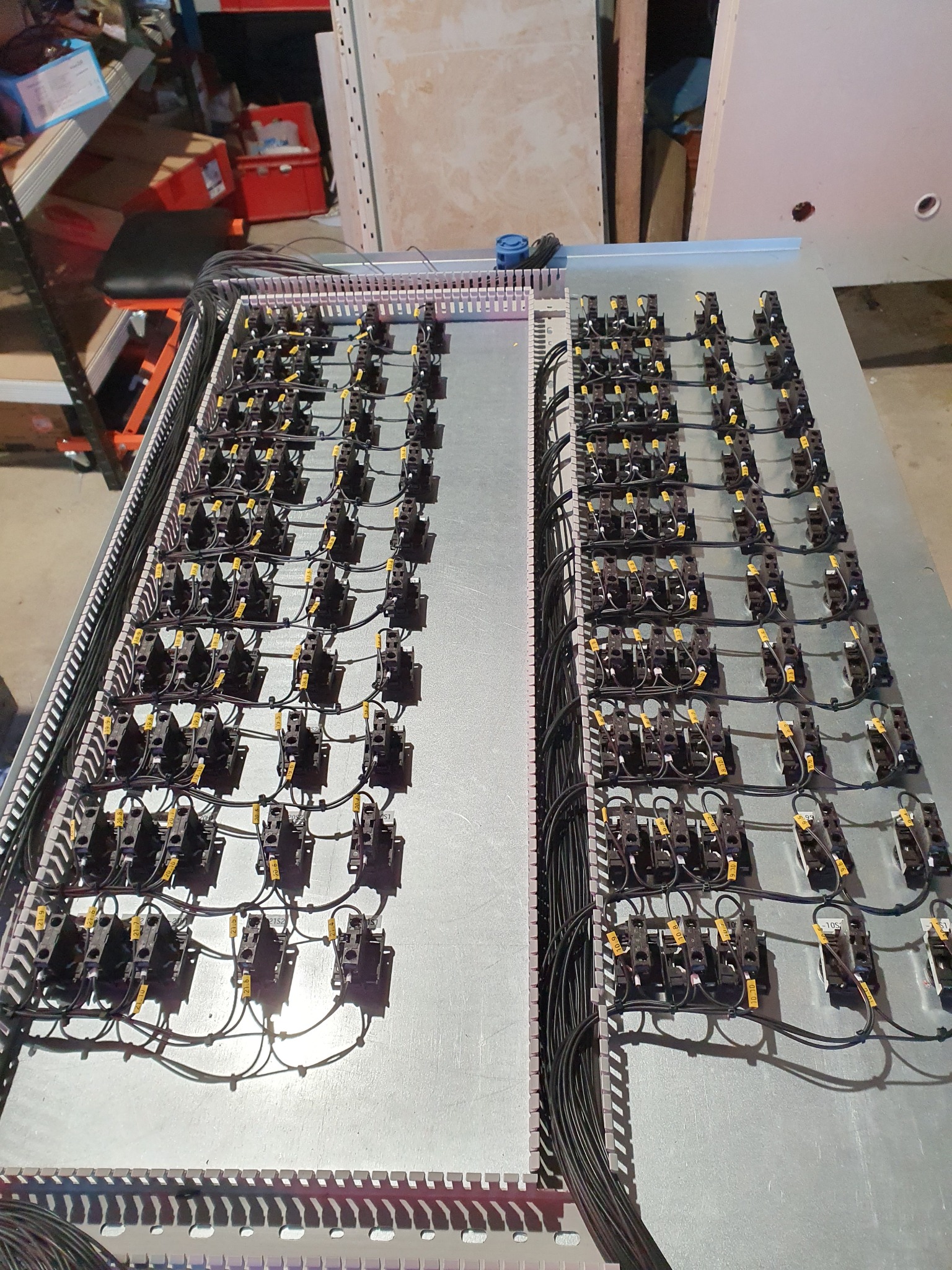 Rozbudowana szafa sterownicza z licznymi przekaźnikami i okablowaniem, widoczne oznaczenia na przewodach, w tle elementy wyposażenia warsztatu.