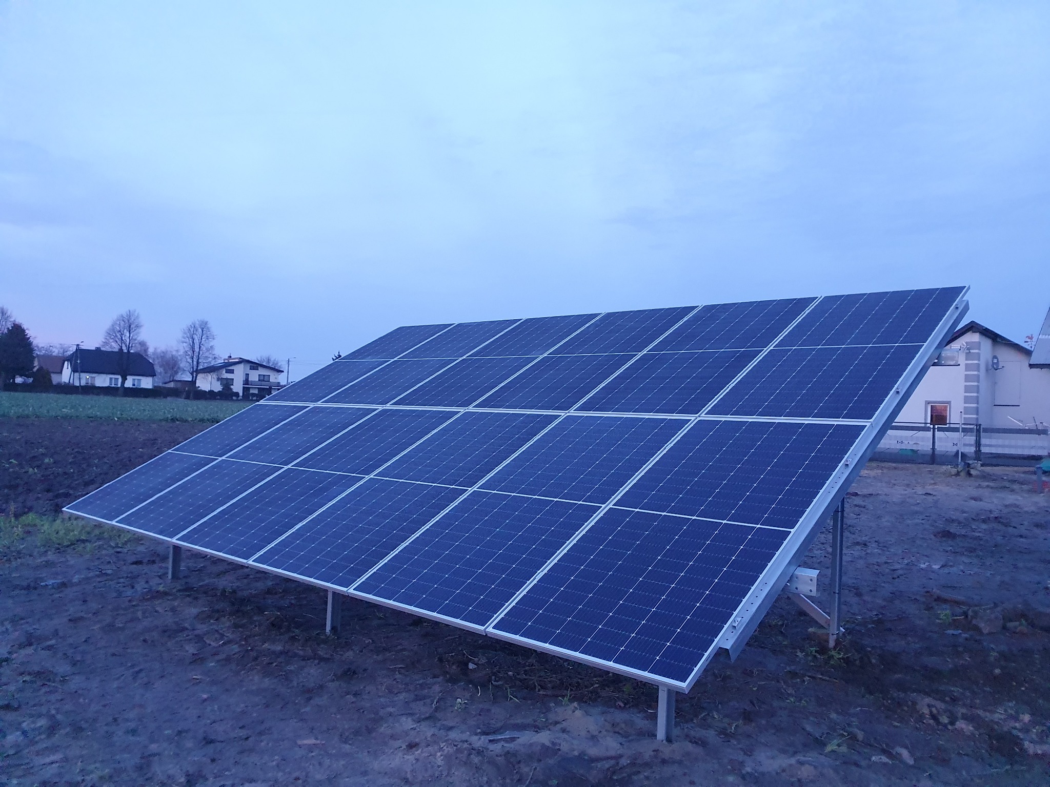 Instalacja paneli fotowoltaicznych na gruncie, widok na rzędy ciemnoniebieskich paneli, tło: domy jednorodzinne i pochmurne niebo.