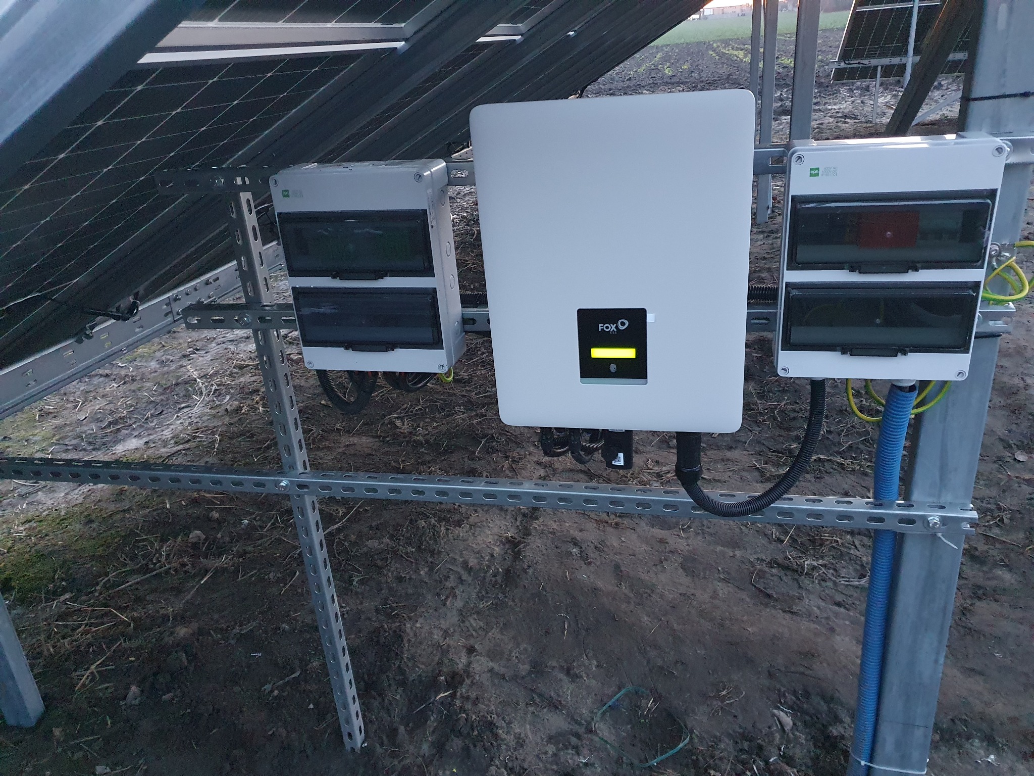 Instalacja paneli słonecznych: widok na inwerter FOX ESS z żółtym wskaźnikiem, skrzynki przyłączeniowe i metalową konstrukcję mocującą na tle ziemi.