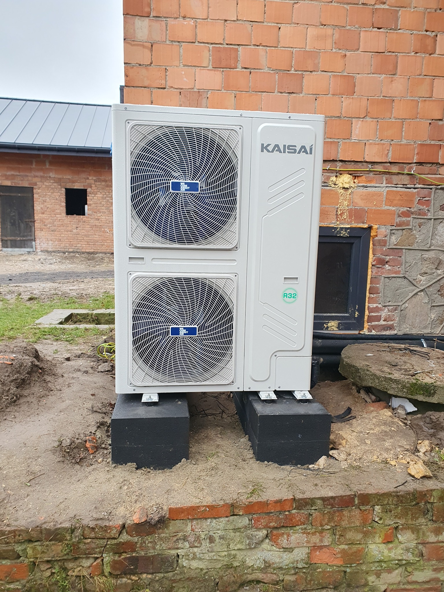 Zewnętrzna jednostka pompy ciepła Kaisai, zamontowana na czarnych blokach fundamentowych przy ceglanej ścianie budynku, widoczny certyfikat R32 i instalacja elektryczna.