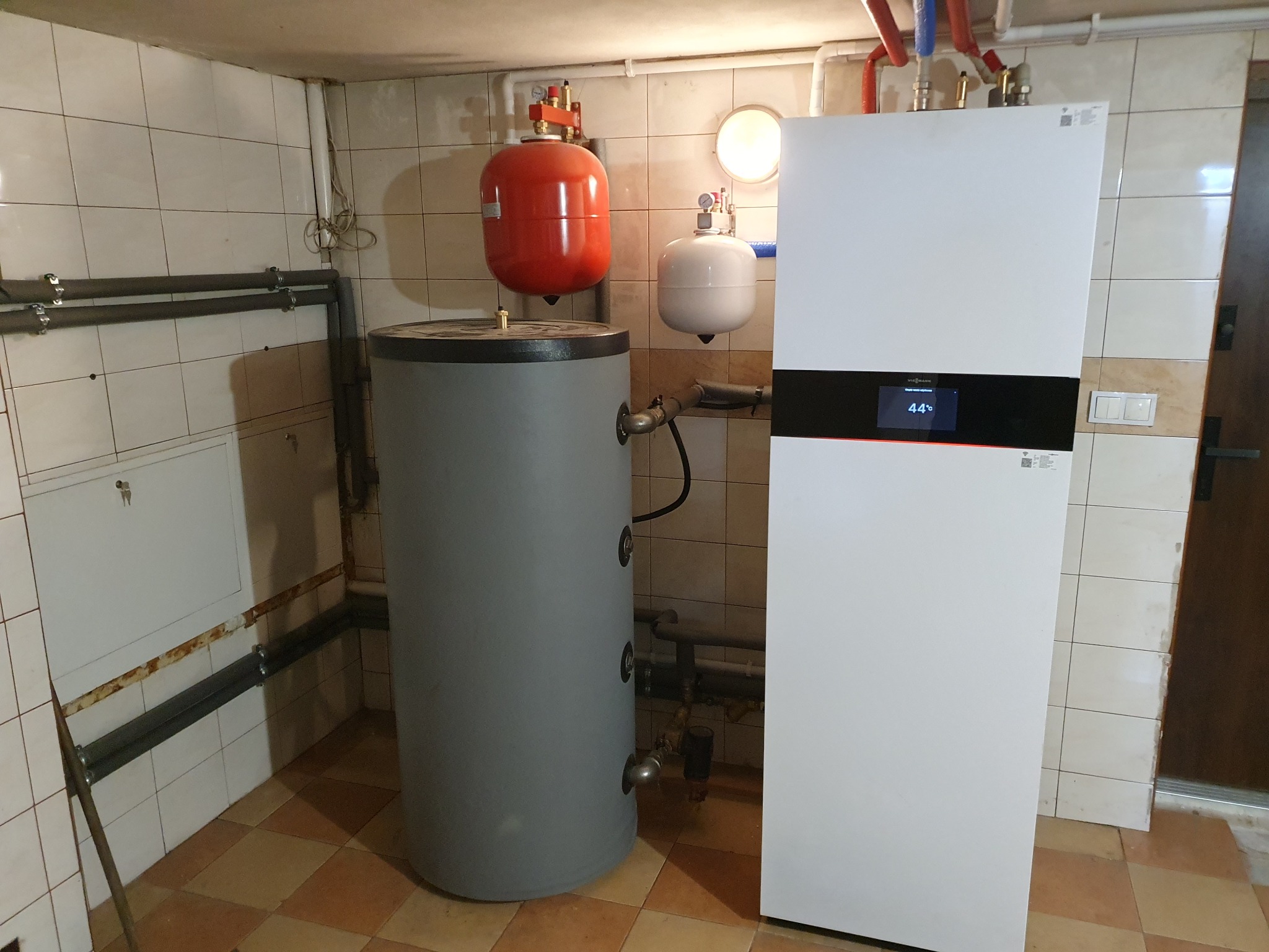 Biała pompa ciepła Viessmann obok szarego zbiornika akumulacyjnego w kotłowni wyłożonej kafelkami, widoczne rury instalacyjne.