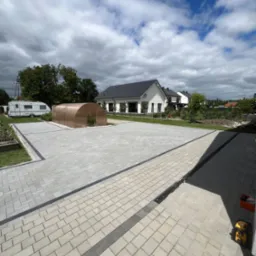 Jasnoszara kostka brukowa ułożona na dużej powierzchni przed domem z widoczną przyczepą kempingową i tunelem foliowym, z odpływem liniowym wzdłuż krawędzi.