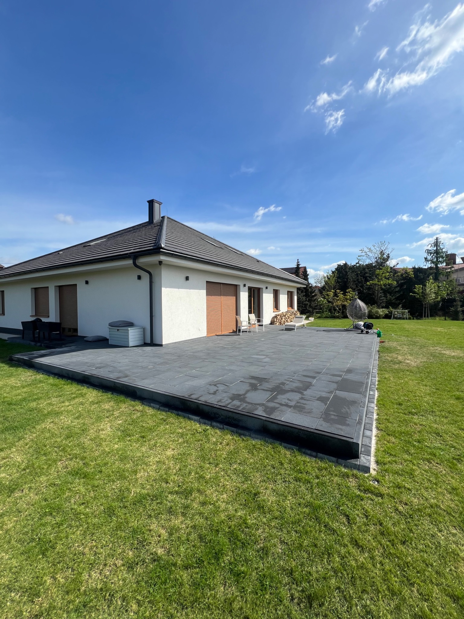 Nowoczesny taras z ciemnoszarej kostki brukowej przylegający do białego domu z brązowymi roletami, widok na zadbany trawnik i elementy wyposażenia ogrodu, takie jak wiszące krzesło i ułożone drewno.