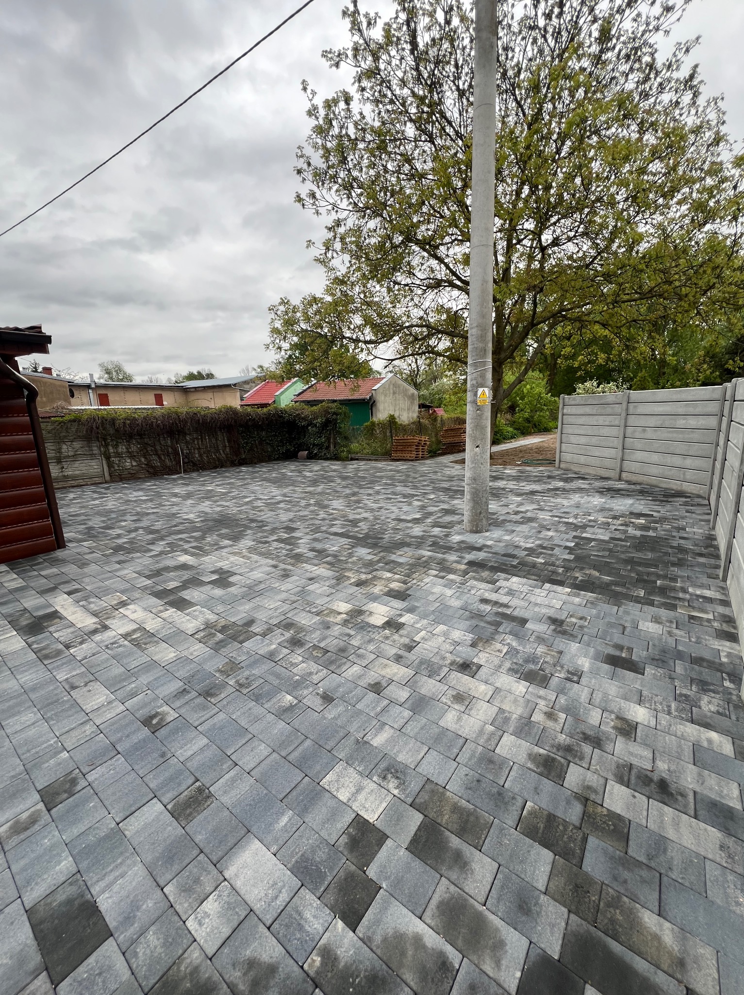 Ułożona szara kostka brukowa na posesji z betonowym ogrodzeniem i słupem energetycznym pośrodku, widok na budynki w tle.