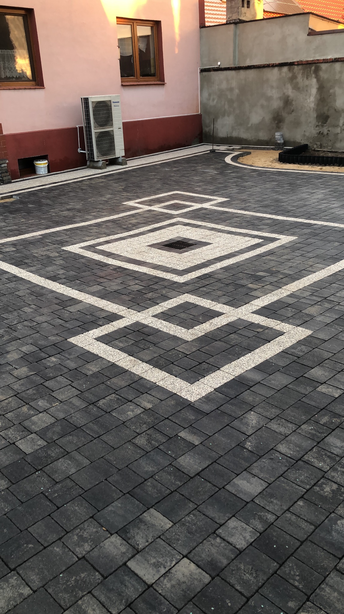 Ułożona kostka brukowa w geometryczny wzór z ciemnoszarej kostki z jasnymi wstawkami, widoczny budynek mieszkalny z klimatyzatorem na ścianie i przygotowane podłoże pod nasadzenia