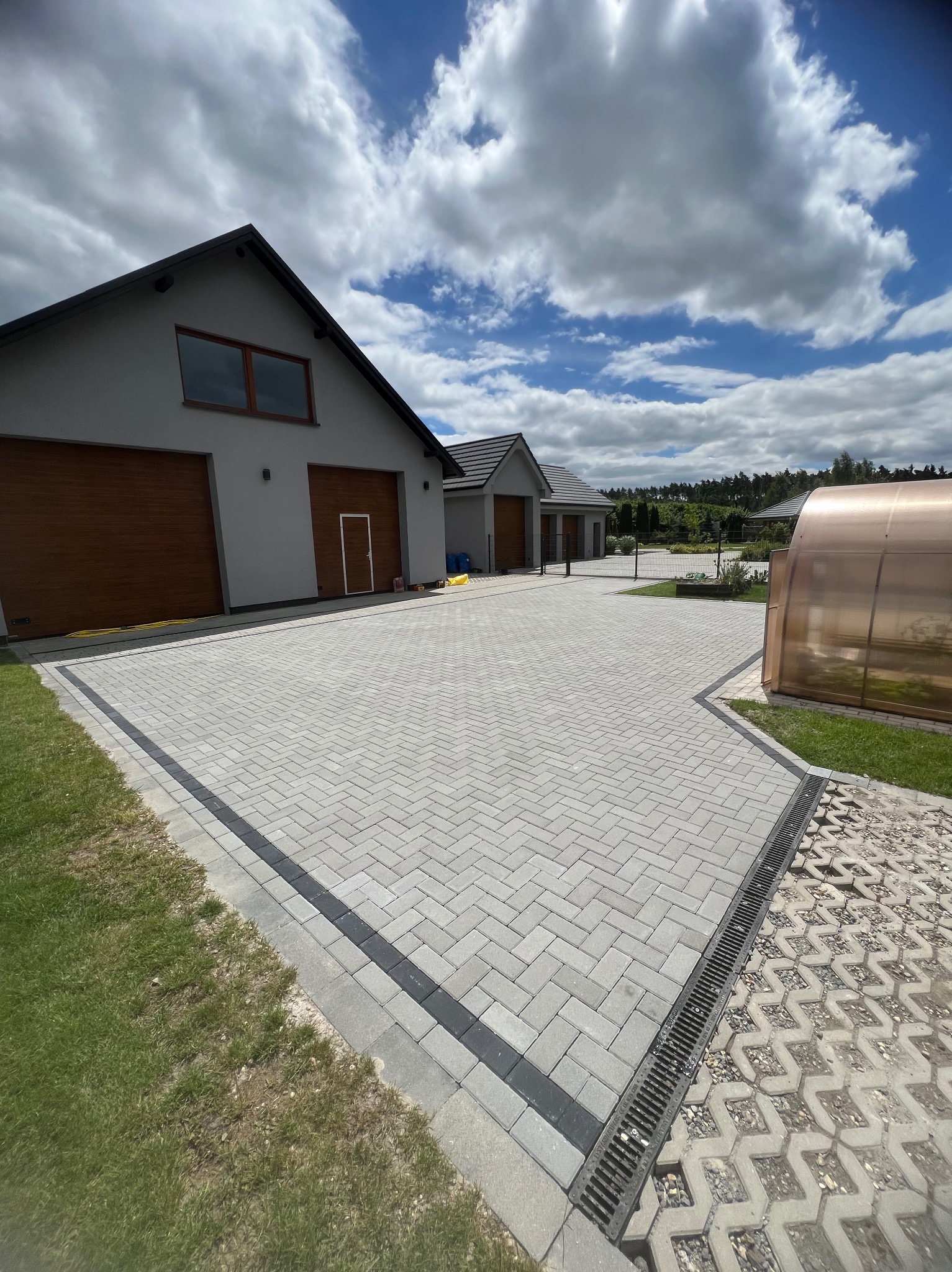 Ułożona kostka brukowa w jodełkę na podjeździe przed domem z garażami, obrzeża z ciemnej kostki, widoczny odpływ liniowy i fragment podjazdu z ażurów, w tle zabudowa jednorodzinna i błękitne niebo...