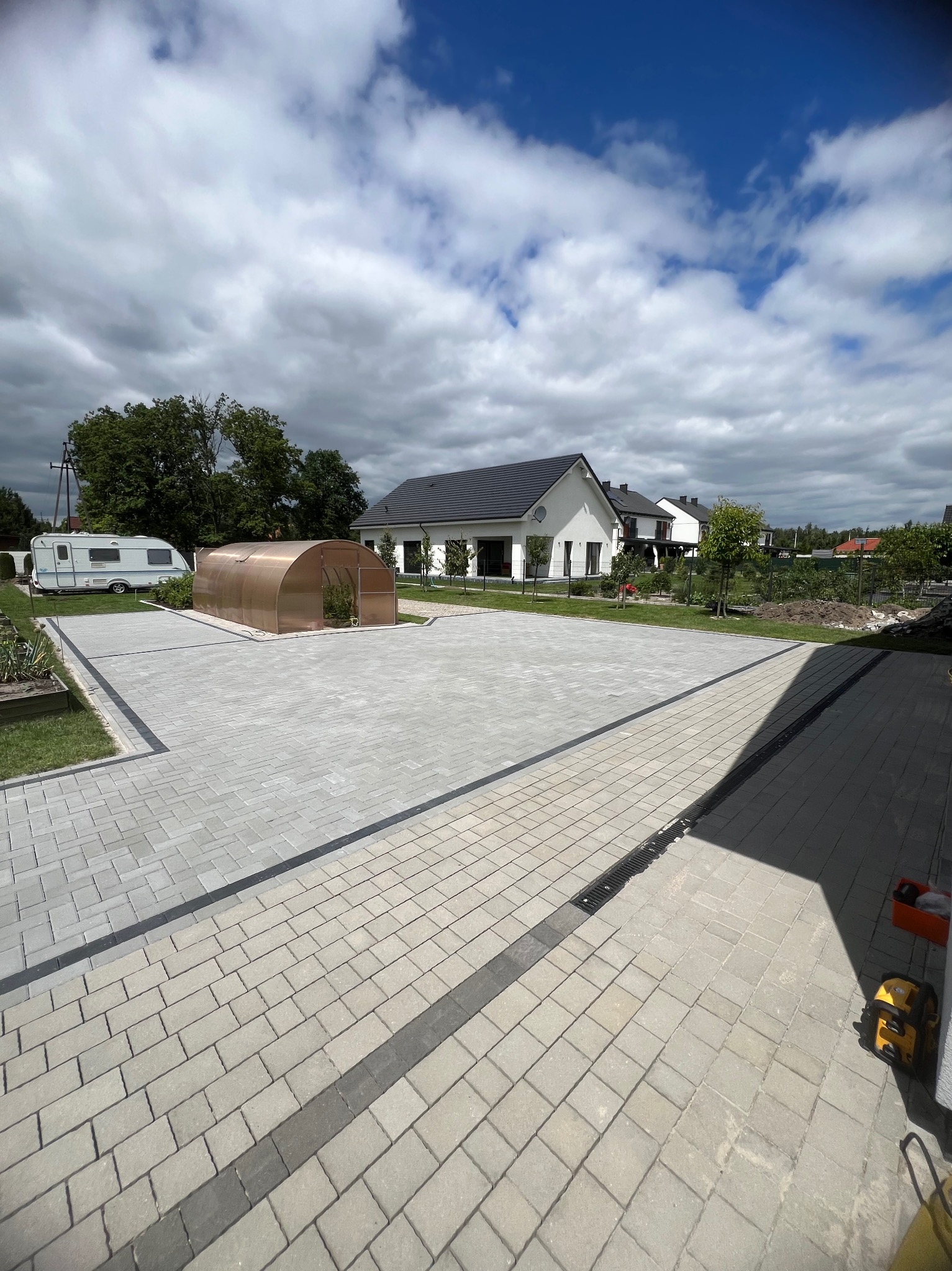 Jasnoszara kostka brukowa ułożona na dużej powierzchni przed domem z widoczną przyczepą kempingową i tunelem foliowym, z odpływem liniowym wzdłuż krawędzi.