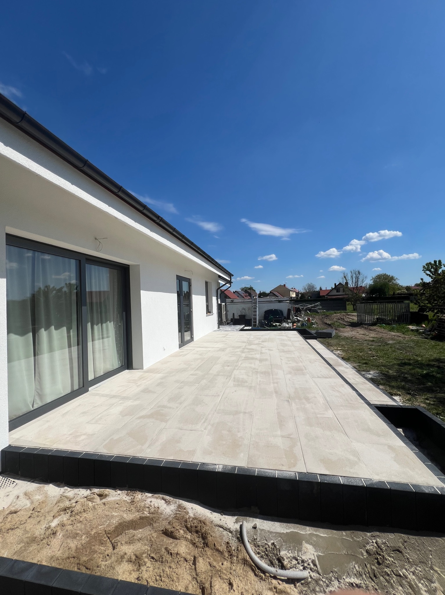 Ułożona jasna kostka brukowa tworząca taras przy nowoczesnym białym domu z dużymi przesuwnymi drzwiami, widoczne czarne obrzeża i przygotowany teren wokół.