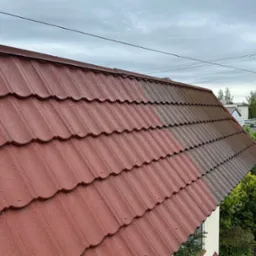 Częściowo wyczyszczony, czerwono-brązowy dach z blachodachówki modułowej, widoczne różnice w kolorze między czystą a zabrudzoną powierzchnią, pochmurne niebo w tle.