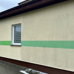 Pod Ciśnieniem - Fragment elewacji budynku z widoczną różnicą w czystości po umyciu fragmentu ściany, z zielonym pasem na górze, szarą ścianą poniżej i widocznym oknem z żaluzjami.
